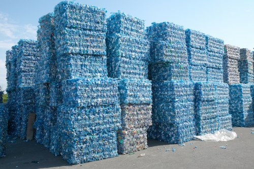 A large outdoor storage area features multiple tall stacks of tightly bound blue plastic water bottles, arranged in rectangular blocks on a paved surface. The bottles are compressed and secured with clear plastic wrapping, creating uniform, stacked formations that reach several meters high. In the background, a clear blue sky and some scattered debris, including a partially visible white plastic sheet, are visible on the ground nearby. The scene suggests a site for bulk waste collection or recycling preparation by a rubbish removal service, consistent with the context of Commercial Waste Carshalton offering specialist waste management solutions in the local area. The overall environment appears clean, with the stacks systematically organized, emphasizing a focus on large-scale disposal or processing of plastic waste material.