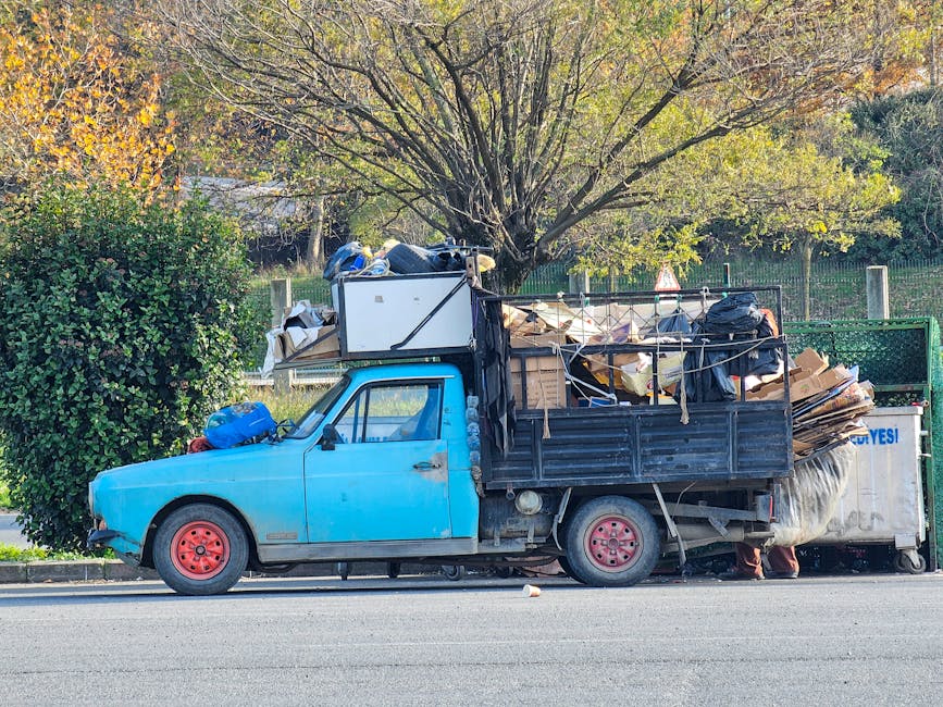 A small, old blue flatbed truck parked along a street, loaded with miscellaneous rubbish including large cardboard boxes, plastic bags, and wooden pallets. The truck's body shows signs of wear with fa