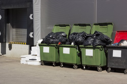 A row of four green wheelie bins with open lids, filled with black rubbish bags placed on a concrete surface outside a commercial building, with three white bricks stacked nearby on the left and a red recycling container partially visible on the right. The background features a grey industrial roller shutter door and grey wall panels, indicating a commercial or industrial waste disposal area in Carshalton. The scene is well-lit, with natural daylight illuminating the bins and surroundings, exemplifying typical refuse collection settings managed by companies like Commercial Waste Carshalton, serving local businesses and waste management needs in the area.